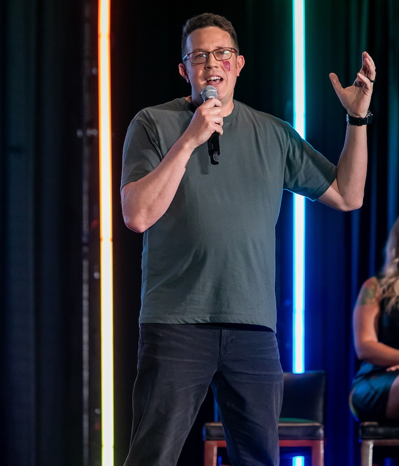 Pete McPherson on stage with a microphone in front of neon light pillars.